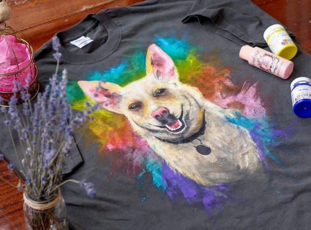 Hand-painted T-shirt with a colorful portrait of a dog, surrounded by paint bottles and dried lavender