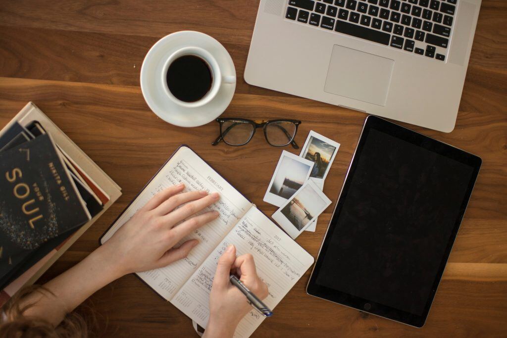 Person writing in a notebook on a desk with a laptop, coffee, and photos.