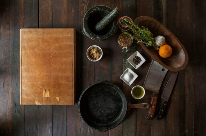 Overhead view of a rustic kitchen setup with a cutting board, cast iron pan, spices, herbs, mortar and pestle, and knives on a wooden surface.