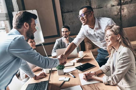 Two male employees shaking hands in the office as other smiling employees watch, capturing a 'welcome to the team' moment