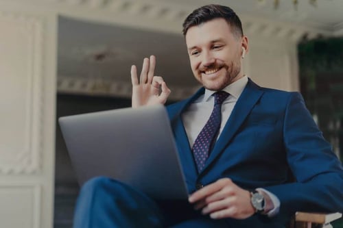 Happy successful businessman in blue navy suit showing ok sign in luxury office during an online video call on laptop, expressing 'thank you, boss'